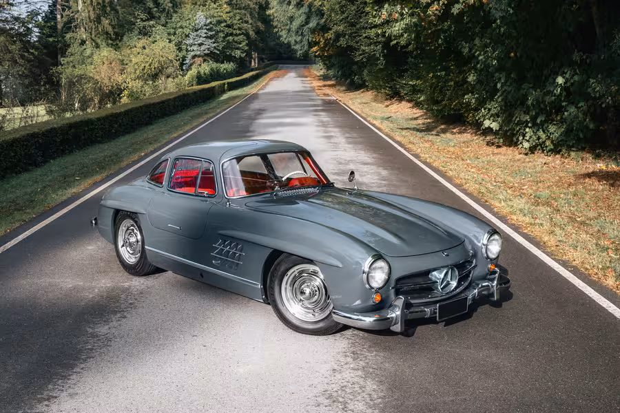 Šedý Mercedes-Benz 300 SL Gullwing z roku 1955 na silnici lemované stromy