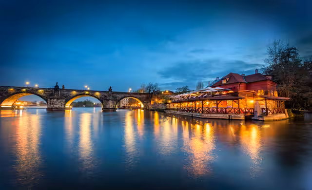 Osvětlená restaurace Kampa Park u Vltavy a nasvícený Karlův most po setmění