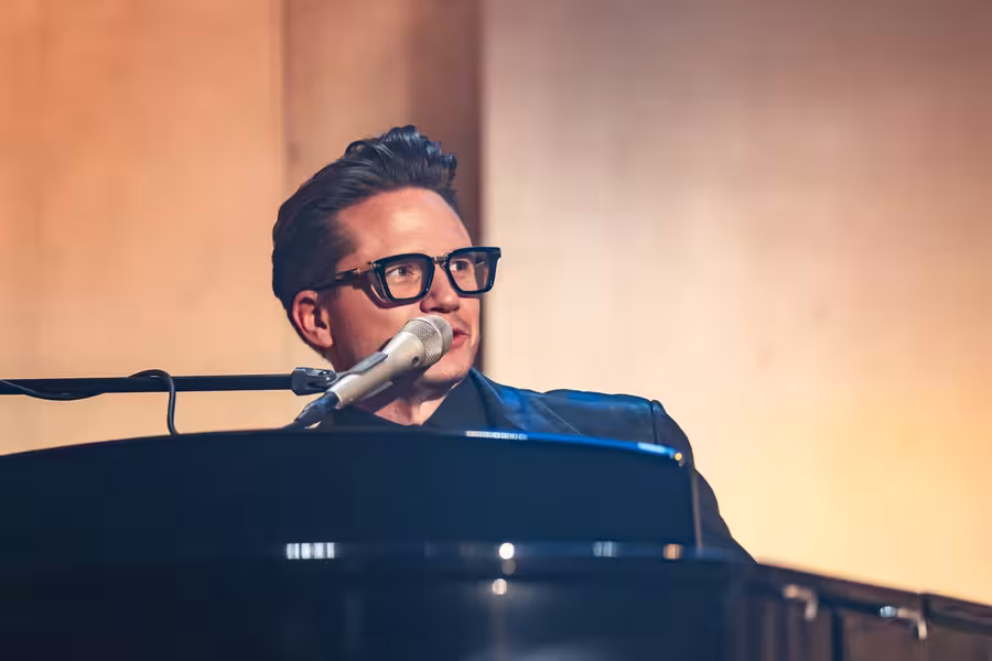 Ondřej Gregor Brzobohatý v brýlích zpívá do mikrofonu za klavírem