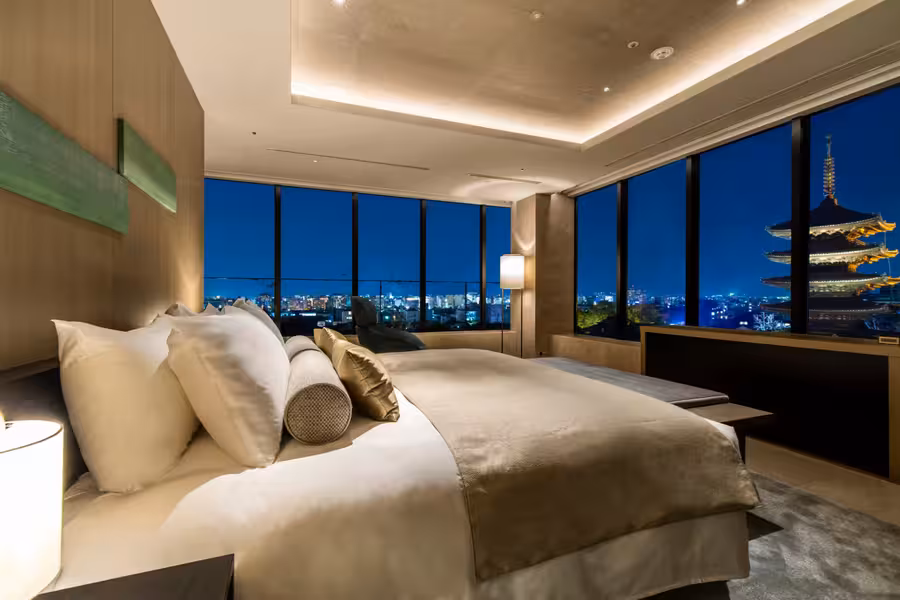 Postel a křeslo v apartmá Panoramic Suite s večerním výhledem na pagodu