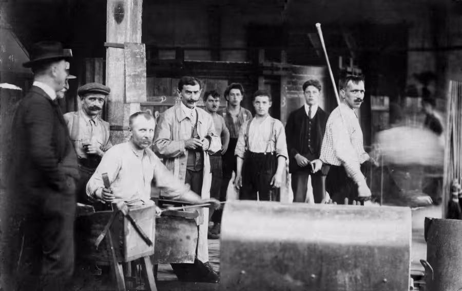 Historický snímek pracovníků ve sklářské hale, s nářadím a pracovním vybavením v popředí