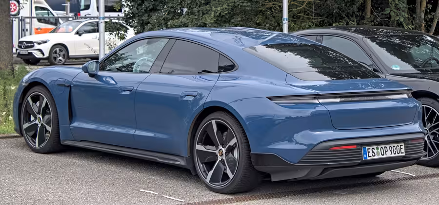 Modré elektrické Porsche Taycan fotografované zezadu z levé strany na parkovišti mezi dalšími automobily