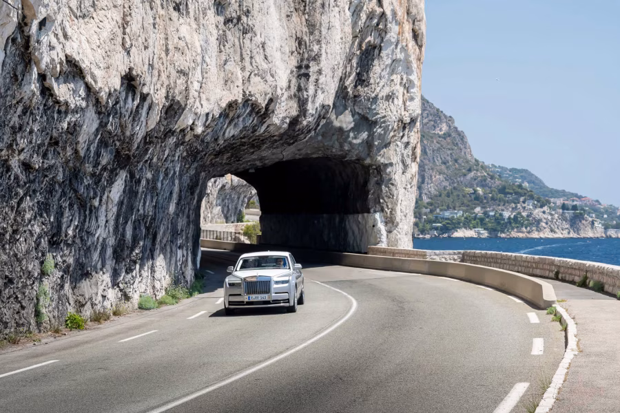 Bílý Rolls-Royce Phantom Series II projíždí po pobřežní silnici u skalního tunelu