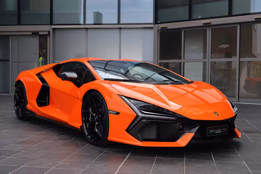 Oranžový Lamborghini Revuelto fotografovaný z nízkého předního tříčtvrtečního pohledu před moderní prosklenou budovou