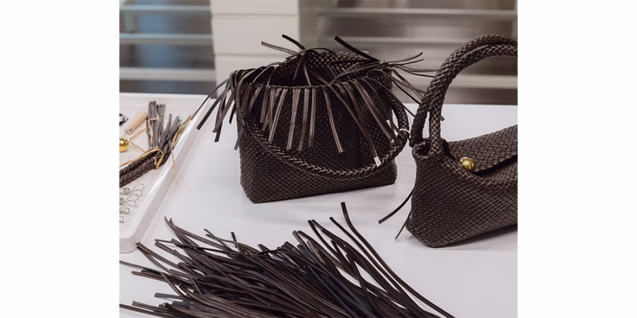 Dvě tmavé pletené kabelky stojí na pracovním stole v ateliéru Bottega Veneta mezi připravenými koženými pásky a nástroji