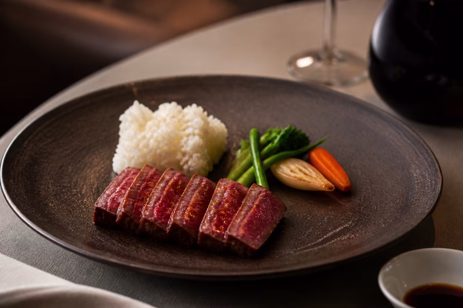 Plátky opečeného Wagyu, kopeček rýže a zelenina na tmavém keramickém talíři.